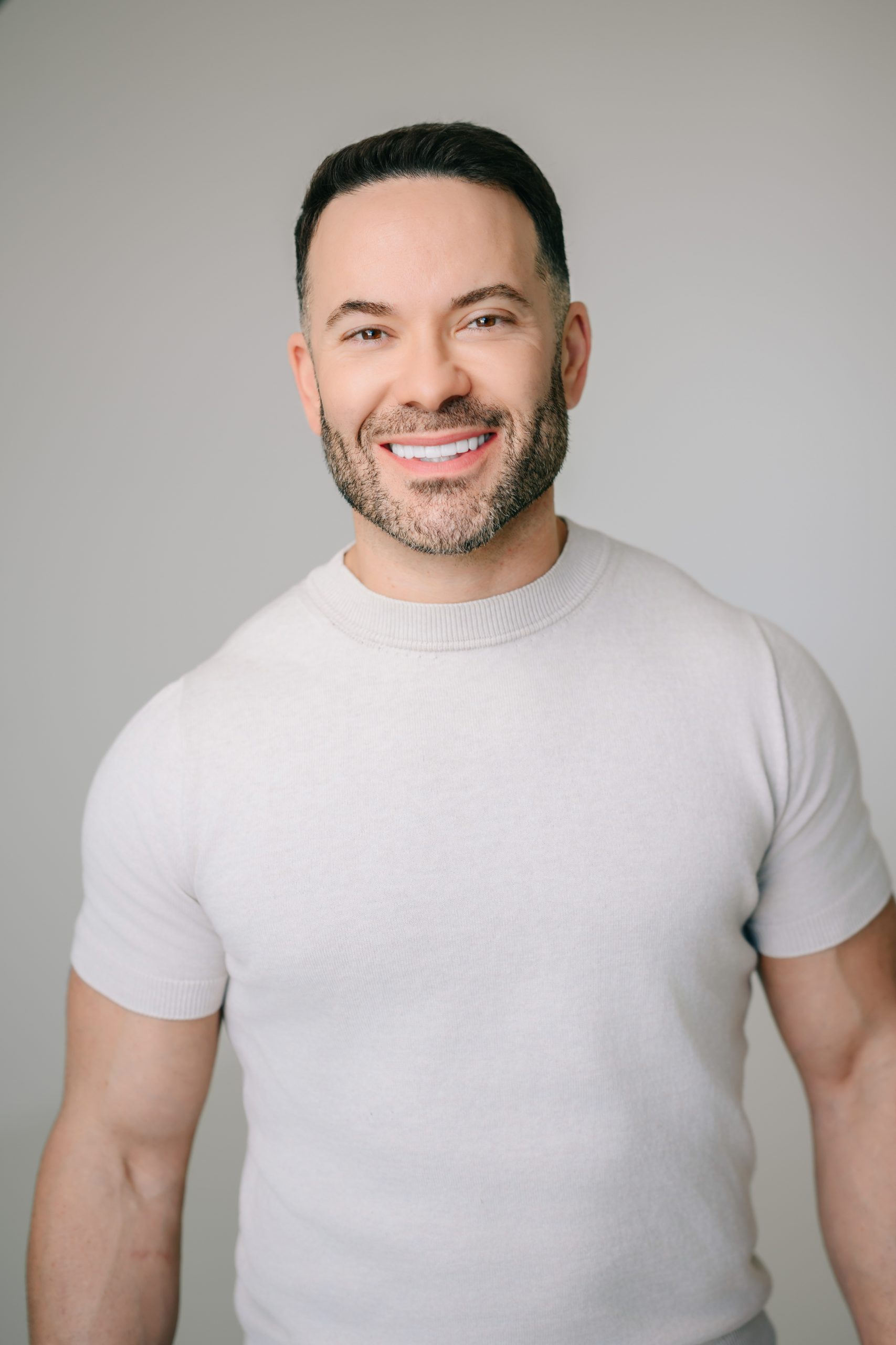 Smiling man with short dark hair and beard wearing a light beige short-sleeve sweater, standing against a soft gray background.