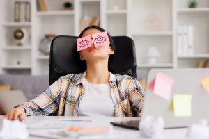 Woman slumped back in her office chair with pink sticky notes drawn with eyes placed over her closed eyes, pretending to be awake at her desk with a laptop and scattered papers