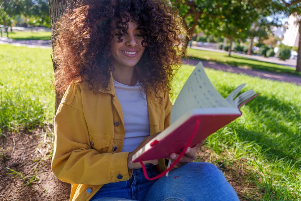 Smiling young woman with curly hair holding a journal in a park, sitting on the grass on a sunny day