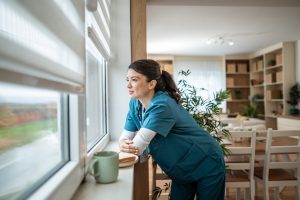 Medical professional standing by a window, looking outside taking a break but feeling calm and contented, not being hard on herself.