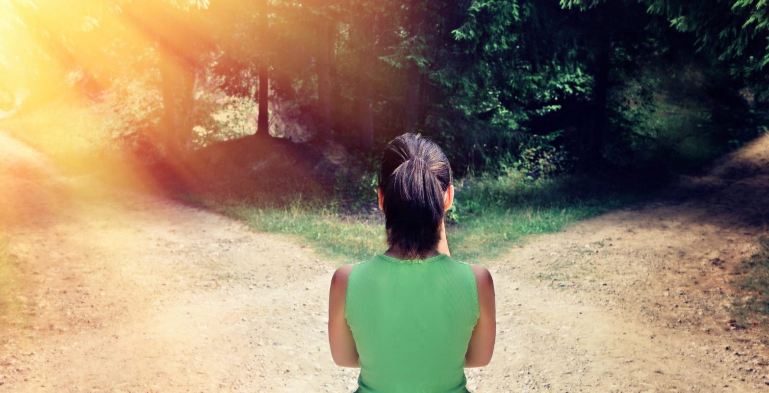 Woman at a forked road, trying to make a choice. One path is bathed in sunlight, the other in shadow.