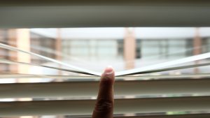 A finger pulls down narrow venetian blinds, revealing a gap of gentle sunlight streaming through, casting soft, intricate shadows and light patterns that evoke a warm, peaceful, and intimate indoor mood.