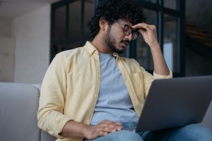 south Asian man in glasses with laptop holding his head, overthinking, self-critical.
