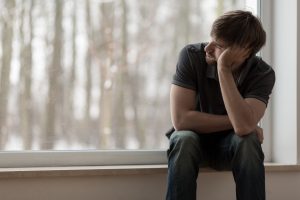 Young man sitting near window with unfocused tall trees behind him, feeling depressed 