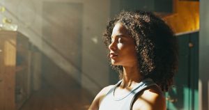 Woman with eyes closed, face turned toward natural light streaming through a window, in a quiet moment of stillness.