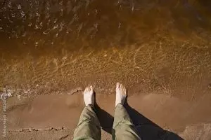 Person standing barefoot at the edge of shallow water, waves lightly touching their feet.