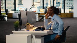 Woman sitting at a desk in an office, leaning forward with her hand over her mouth while looking at dual computer screens, appearing mentally fatigued and overwhelmed.