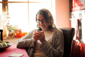 Woman sitting in an armchair at home, holding a warm mug with both hands, looking down with a quiet, reflective expression.