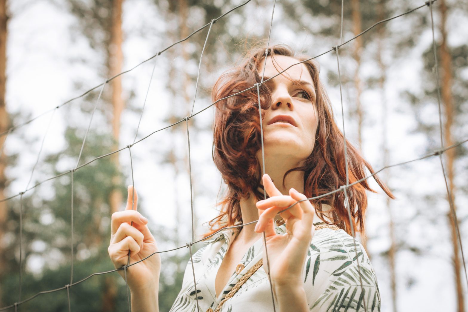 Woman standing behind a wire fence in a forest, looking outward with a thoughtful, emotionally distant expression, symbolizing feeling stuck in old patterns or beliefs.