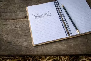 An open spiral notebook rests on a rustic wooden table beside a pencil. The word “impossible” is handwritten on the page and crossed out with dark lines. The image symbolizes questioning long-held beliefs, reframing self-limiting thoughts, and creating space for new possibilities.