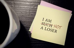 A yellow sticky note on a dark wooden surface reads “I am such a loser,” with the word “such” crossed out and replaced with “not” in red ink. A coffee mug sits blurred in the foreground. The image represents questioning harsh self-talk and rewriting long-held negative beliefs about oneself.