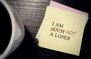 A yellow sticky note on a dark wooden surface reads “I am such a loser,” with the word “such” crossed out and replaced with “not” in red ink. A coffee mug sits blurred in the foreground. The image represents questioning harsh self-talk and rewriting long-held negative beliefs about oneself.