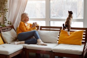Woman relaxing at home on a couch with a cup of tea, watching her cat look out the window on a winter morning