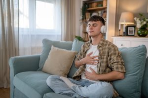Person sitting cross-legged on a couch with headphones on and eyes closed, one hand on their chest and one on their stomach in a calm home setting.