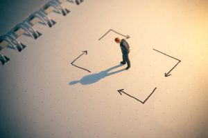 Miniature person standing in the center of hand-drawn arrows pointing in different directions, symbolizing overthinking, mental loops, and difficulty making decisions.