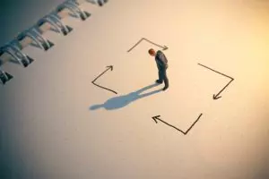 Miniature person standing in the center of hand-drawn arrows pointing in different directions, symbolizing overthinking, mental loops, and difficulty making decisions.