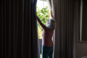 Woman with curly grey hair standing by a window opening dark curtains to reveal bright sunlight and greenery outside.
