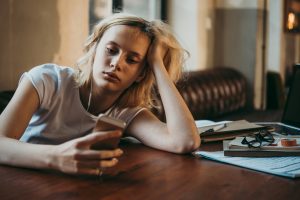 Person sitting at a table looking at a phone while work materials remain untouched, illustrating avoidance behavior and procrastination.