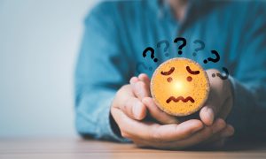 Person holding a worried face icon surrounded by question marks, representing the anxiety, self-doubt, and overthinking that can follow conflict avoidance and self-silencing.