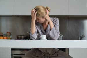 Person sitting alone at a kitchen counter with head in hands, appearing overwhelmed by persistent self-critical thoughts and mental stress, illustrating the constant background noise often associated with functional freeze.