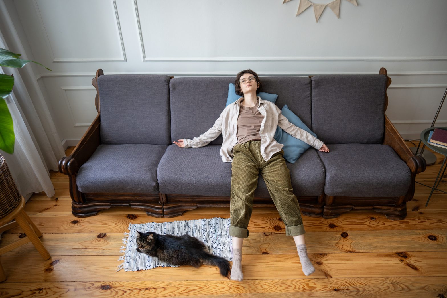 Person sitting slumped on a couch in a bright living room while a cat rests on the floor nearby, illustrating the exhaustion, disconnection, and stuckness often associated with functional freeze.