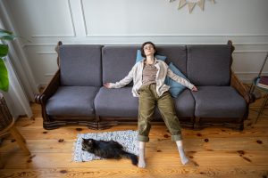 Person sitting slumped on a couch in a bright living room while a cat rests on the floor nearby, illustrating the exhaustion, disconnection, and stuckness often associated with functional freeze.