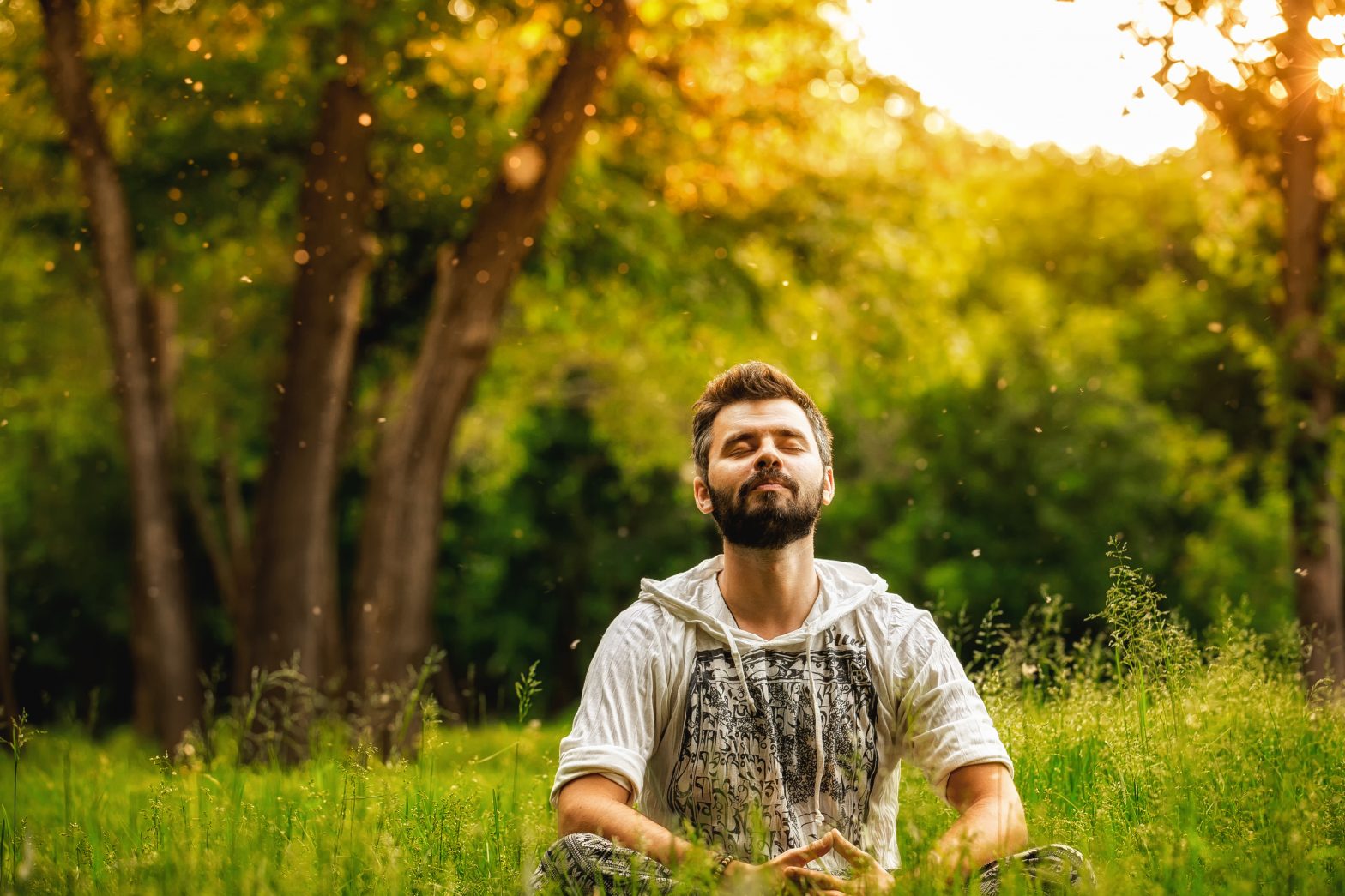 Person sitting cross-legged in a sunlit meadow with eyes closed, surrounded by warm golden light and floating particles, appearing calm and present in the moment.