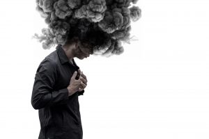 A person stands with their head bowed while a large cloud of dark smoke rises from their head, representing overthinking, emotional overwhelm, and intrusive negative beliefs.