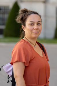 Woman in her 40s outside in orange blouse with her hair up, carrying a small backpack, smiling at the camera with a calm, confident expression, showing she's feeling better and more like herself.