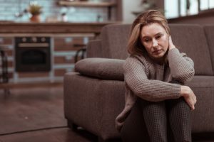 Woman sitting on the floor beside a sofa, looking withdrawn and emotionally distressed. 