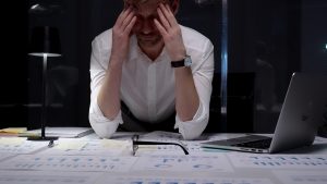 Overwhelmed man holding his head while working late at a desk covered with documents and a laptop, thinking about finding therapy for burnout.