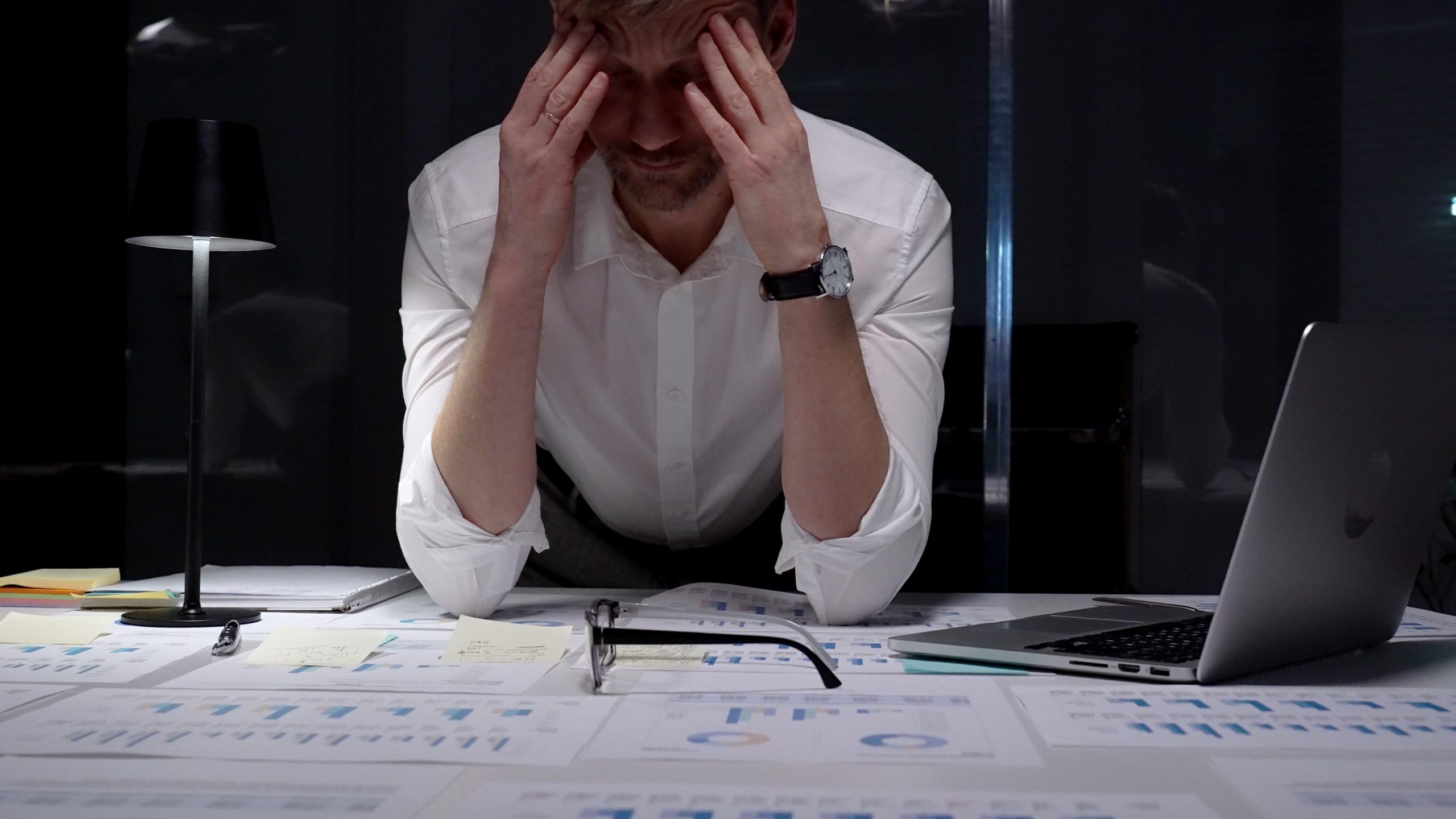 Overwhelmed man holding his head while working late at a desk covered with documents and a laptop, thinking about finding therapy for burnout.