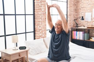 An older man in a brick-walled bedroom stretching after a great nights sleep, getting better rest after regulating the nervous system.