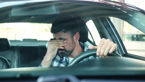 Stressed man sitting behind the steering wheel of a car while covering his eyes with one hand, experiencing functional freeze. 