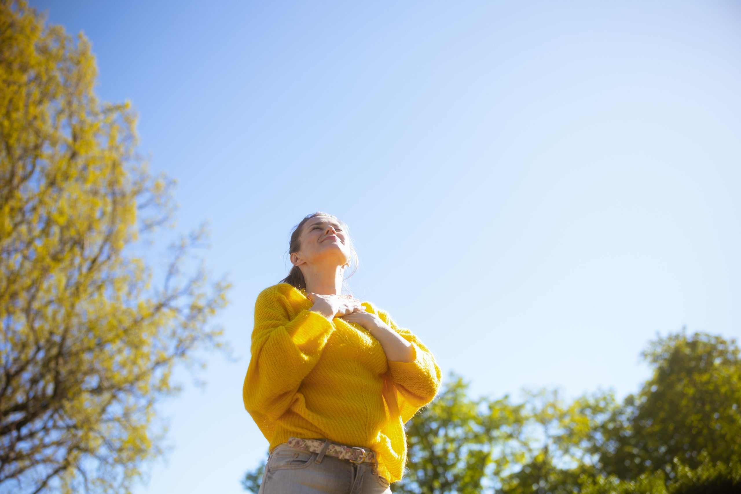 Camera angle looking up at a woman in a yellow sweater taking deep breath with her hands on her sternum. She is outside on a bright day near a tree with yellow fall leaves. Representiing the deep calm and ease brought about by ketamine therapy.