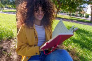 Woman with long curly hair smiling while sitting outside on a sunny lawn near a tree, holding a red journal.