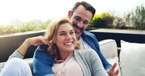 A man with his arms around a woman seated outside on a deck, smiling, laughing, enjoying each other's company, representing potential for more connected relationships with ketamine therapy.