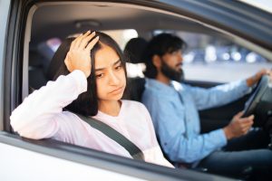 A woman looking out of the passenger side window with head in her hand, detached from her partner who is driving, due to emotional exhaustion.