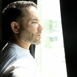 A man in a white t-shirt looks out at a bright sunny day through the space between black curtains, seeming peaceful.